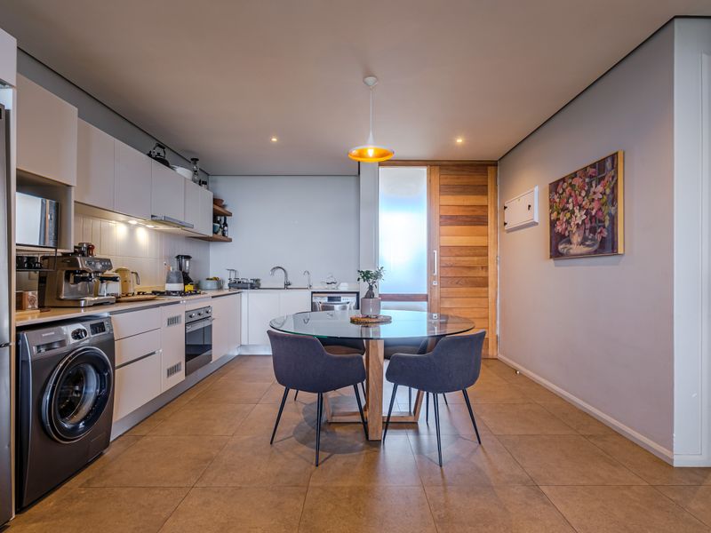 Modern open-plan kitchen and dining area featuring sleek cabinetry, integrated appliances, tiled flooring, and a warm timber door leading to adjacent spaces.