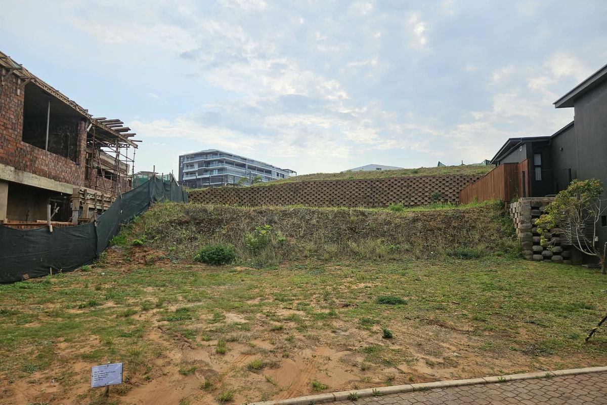 A vacant residential plot featuring a tiered retaining wall, paved access, and surrounding construction activity in a developing neighborhood.