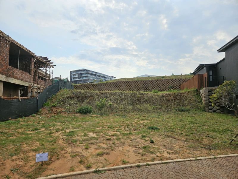 A vacant residential plot featuring a tiered retaining wall, paved access, and surrounding construction activity in a developing neighborhood.
