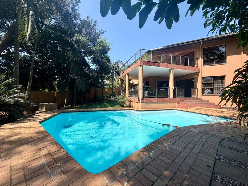 Rear view of the property featuring a swimming pool, brick paved surround, and a raised patio with glass balustrades.