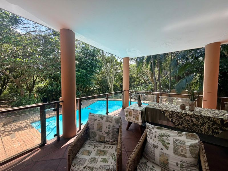 A covered outdoor patio featuring a glass balustrade, overlooking a private swimming pool and lush garden surroundings.