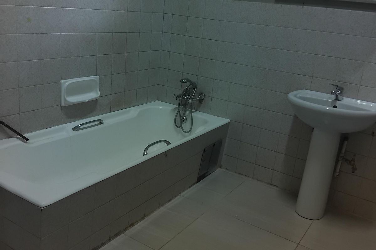 A functional bathroom featuring a bathtub with mixer tap, pedestal basin, and tiled finishes, currently requiring minor repairs to the bath panel and plumbing.
