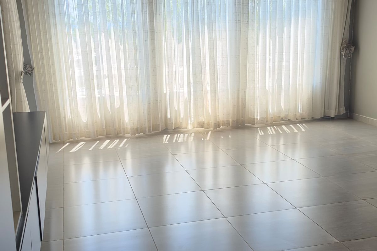 A bright, empty living room featuring large windows with sheer curtains, exposed timber ceiling beams, tiled flooring, and a wall-mounted split-system air conditioner.
