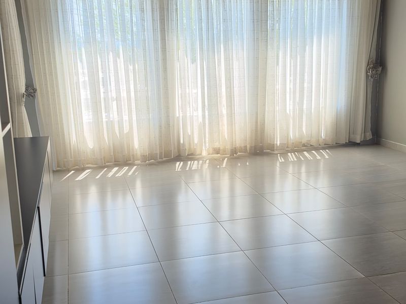 A bright, empty living room featuring large windows with sheer curtains, exposed timber ceiling beams, tiled flooring, and a wall-mounted split-system air conditioner.