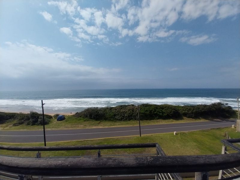 Elevated coastal view overlooking the road, coastal vegetation, and the ocean, framed by a timber balcony railing.