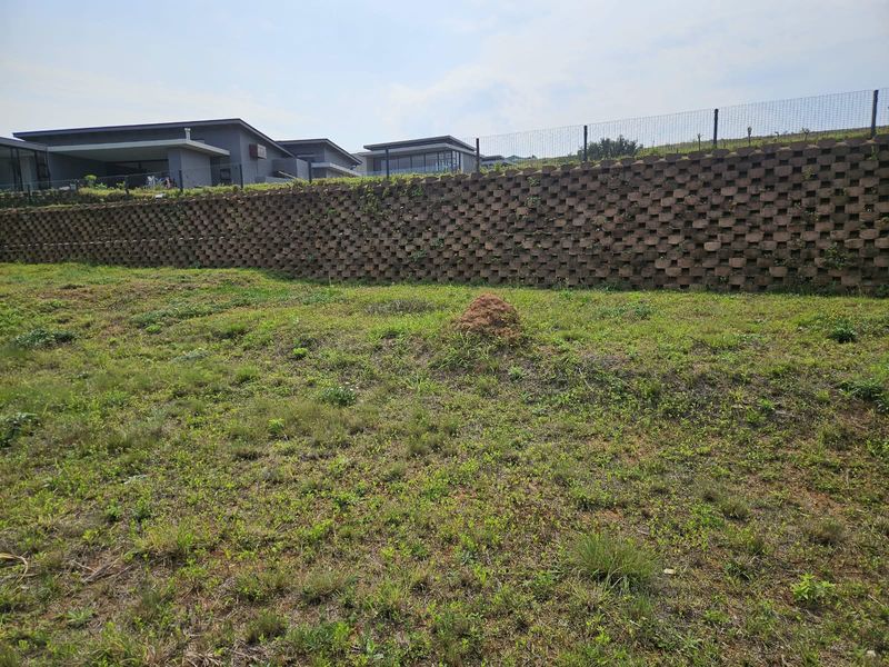 A vacant residential plot featuring a tiered concrete block retaining wall and perimeter wire fencing, set against a backdrop of modern neighboring homes.