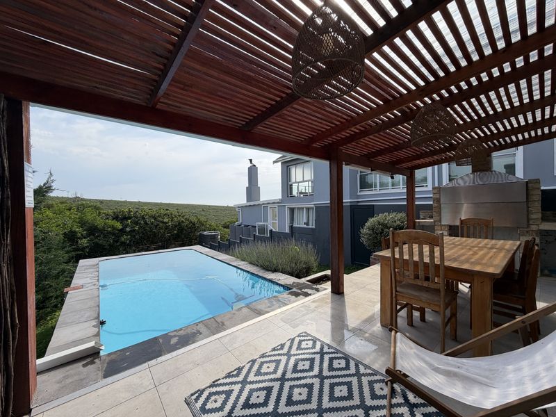 Covered outdoor patio featuring a timber pergola, built-in braai, and a swimming pool overlooking the surrounding landscape.