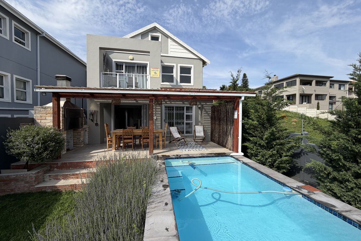 Rear exterior view featuring a swimming pool, a covered patio with a built-in braai, and an upper-level balcony.