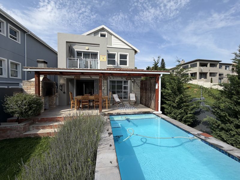 Rear exterior view featuring a swimming pool, a covered patio with a built-in braai, and an upper-level balcony.