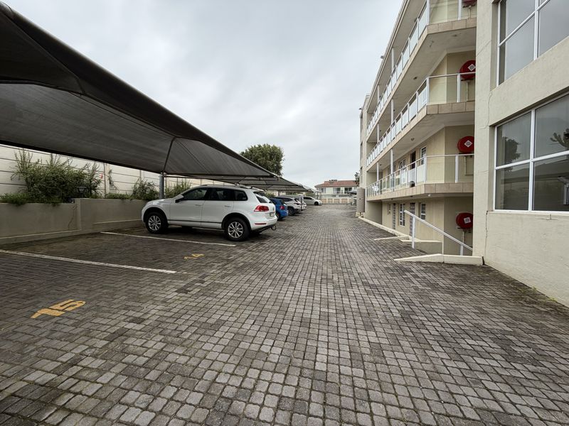 Exterior view of the apartment complex parking area featuring paved driveway, shade cloth carports, and building facade with balcony access.