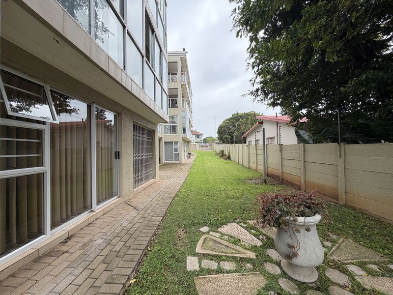 Rear exterior view of the apartment building featuring a paved walkway, lawn area, and secure boundary wall with electric fencing.