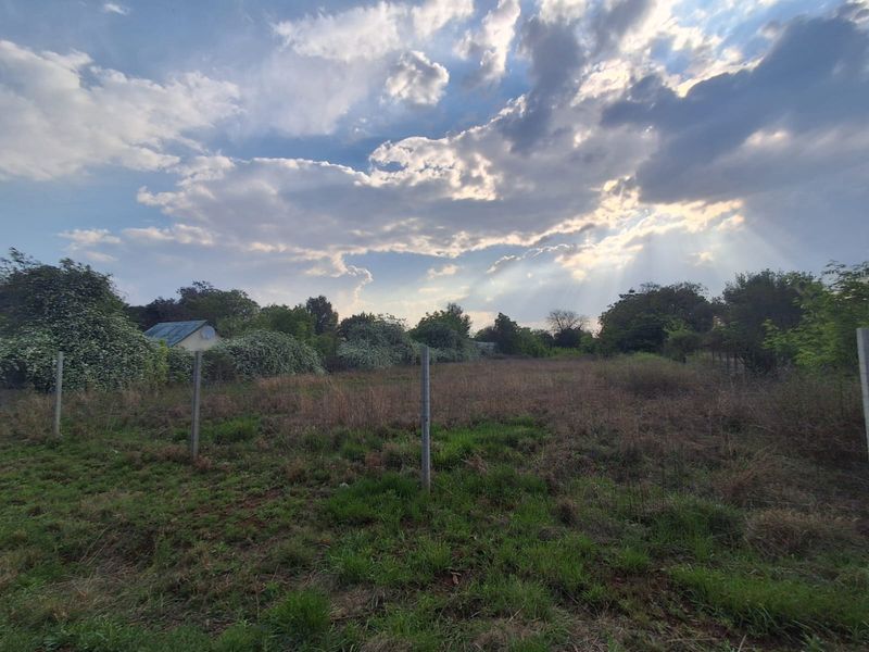A view of a vacant residential plot featuring open grassy land, perimeter fencing, and mature surrounding vegetation under a cloudy sky.