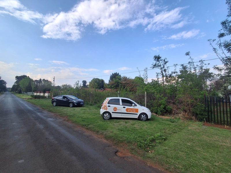 A street-level view of a vacant residential plot featuring a grass verge and wire mesh perimeter fencing.