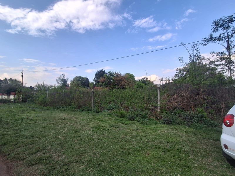 A view of a vacant residential plot featuring grass coverage, overgrown boundary vegetation, and existing wire fencing with concrete posts.