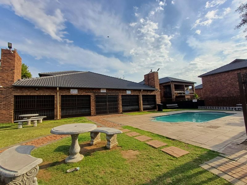 Communal pool area featuring a paved deck, brick building exterior with security gates, and landscaped garden space.