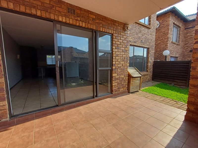 A covered patio area with tiled flooring, featuring a sliding glass door entrance into the apartment and access to a small garden space.