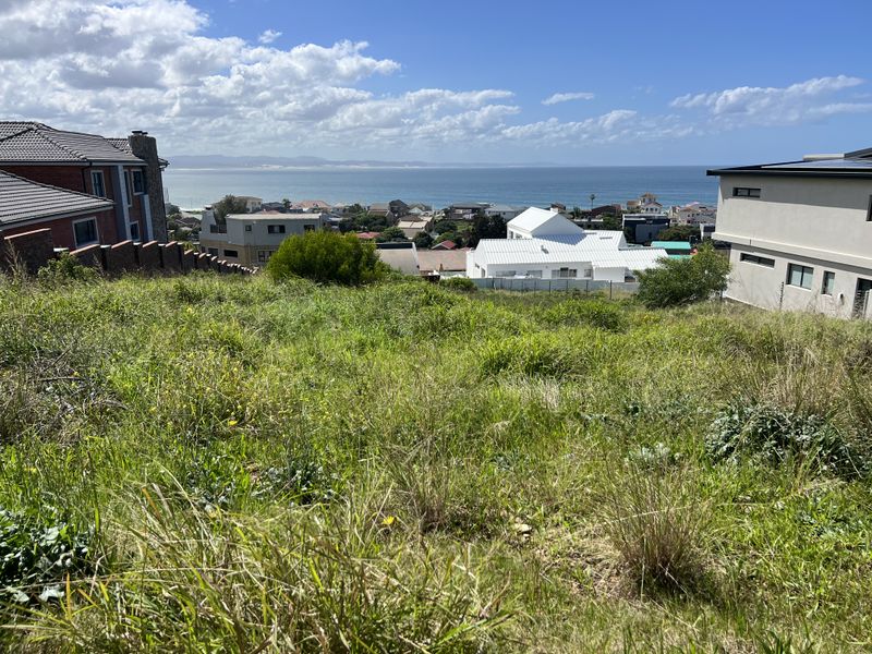A vacant residential plot featuring a sloped landscape with expansive coastal and ocean views, surrounded by established residential properties.