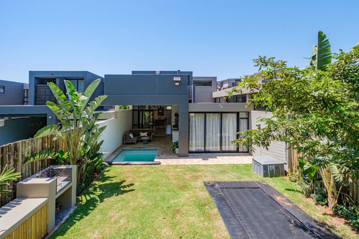 Rear view of a modern home featuring a private swimming pool, paved patio area, built-in braai, and a well-maintained garden.