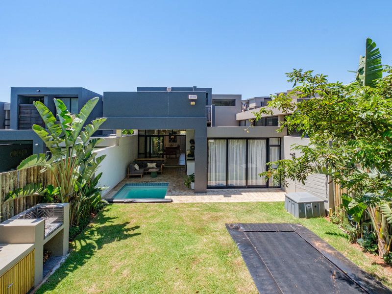 Rear view of a modern home featuring a private swimming pool, paved patio area, built-in braai, and a well-maintained garden.