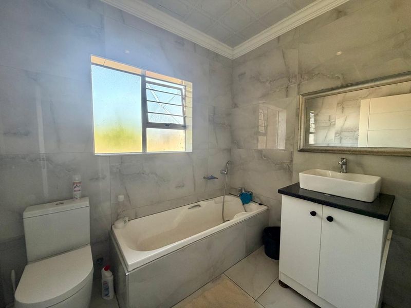A tiled bathroom featuring a built-in bathtub, a vanity cabinet with a vessel sink, and a toilet. The window pane is currently cracked.