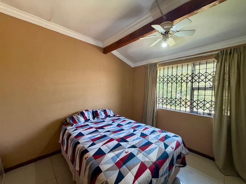 A bedroom featuring tiled flooring, an exposed timber beam, a ceiling fan, and a window fitted with a security gate.