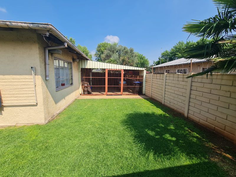 A rear garden area featuring a well-maintained lawn, a concrete block boundary wall with security wire, and a covered patio with a metal security gate.