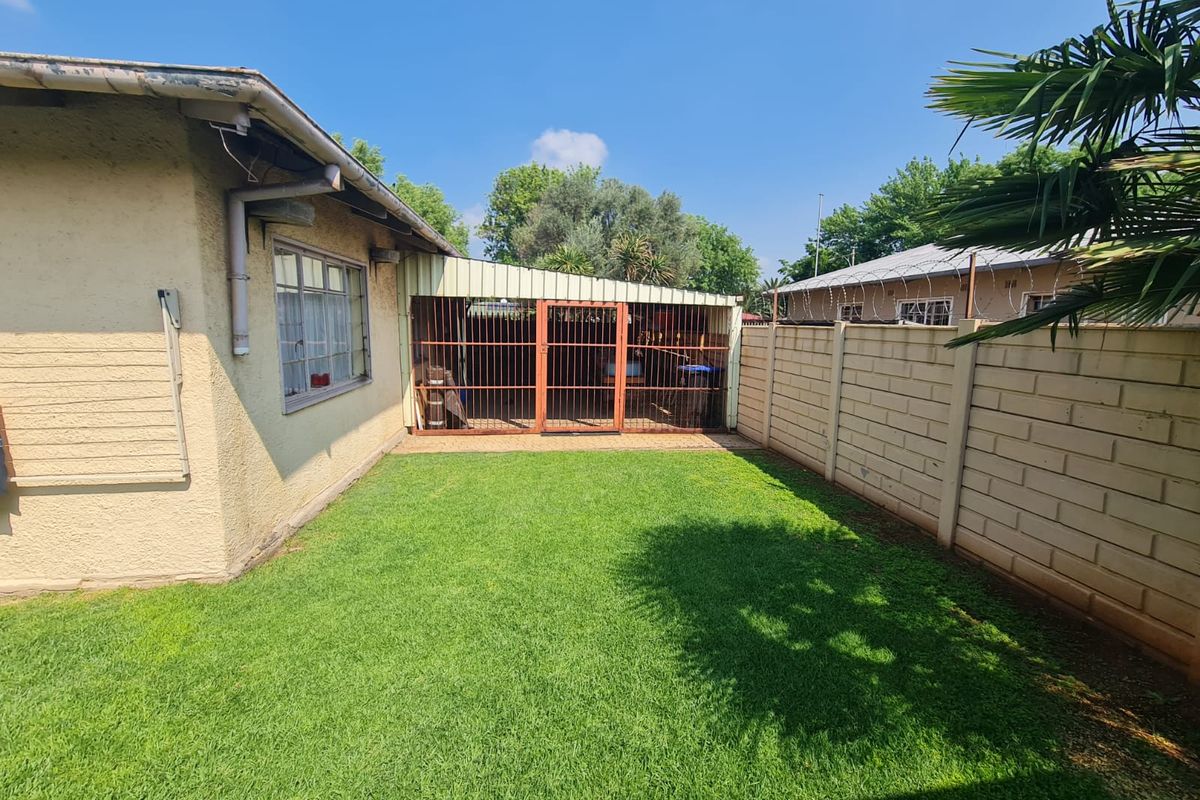 A rear garden area featuring a well-maintained lawn, a concrete block boundary wall with security wire, and a covered patio with a metal security gate.