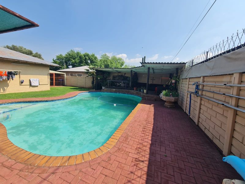 Outdoor pool area featuring a swimming pool, brick paved surround, and a covered patio structure, enclosed by a perimeter wall with security spikes.