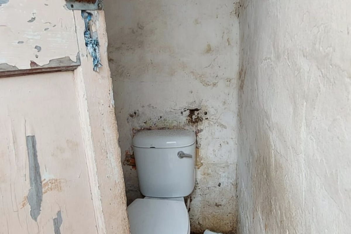 A small, dilapidated outdoor toilet facility requiring significant renovation and repair to the walls, ceiling, and flooring.