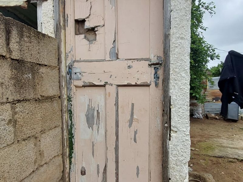 Exterior view of a utility structure featuring a severely weathered and damaged timber door with significant structural deterioration.