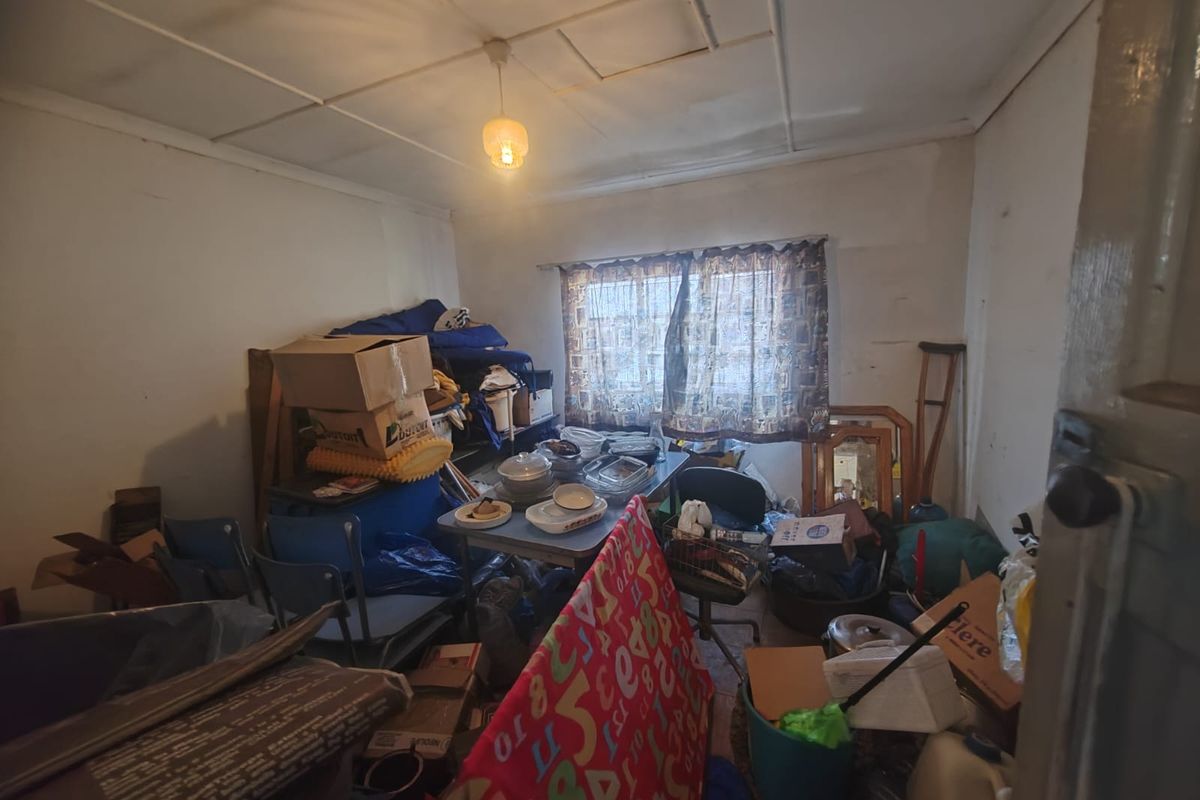 A bedroom space requiring significant renovation, featuring original ceiling tiles and window fixtures, currently cluttered with personal belongings.