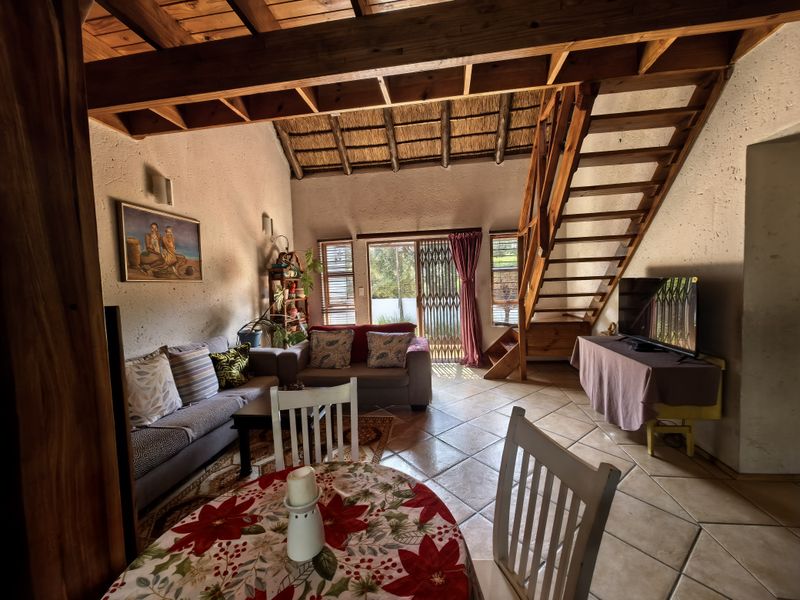 Open-plan living area featuring a double-volume thatched ceiling, exposed timber beams, a wooden staircase, and tiled flooring with direct access to the patio.