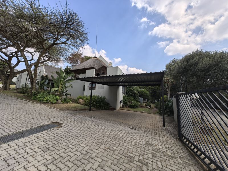 Front exterior view of the townhouse featuring a paved driveway, covered carport, and established garden with a distinctive thatch roof structure.