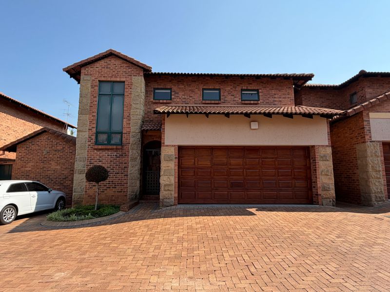 Front elevation of a double-story townhouse featuring a brick and stone facade, tiled roof, and a wooden double garage door with a paved driveway.