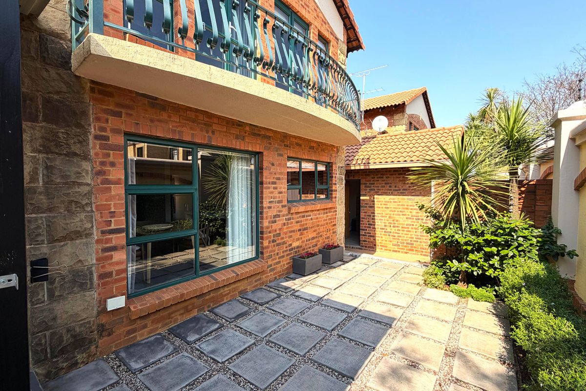 A well-maintained rear exterior view of a brick townhouse featuring a paved patio area and an upper-level balcony with metal railings.