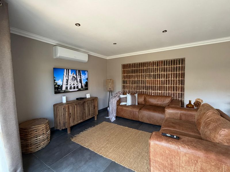 A well-maintained living room featuring a distinctive exposed brick accent wall, modern tiled flooring, recessed lighting, and a wall-mounted air conditioning unit.
