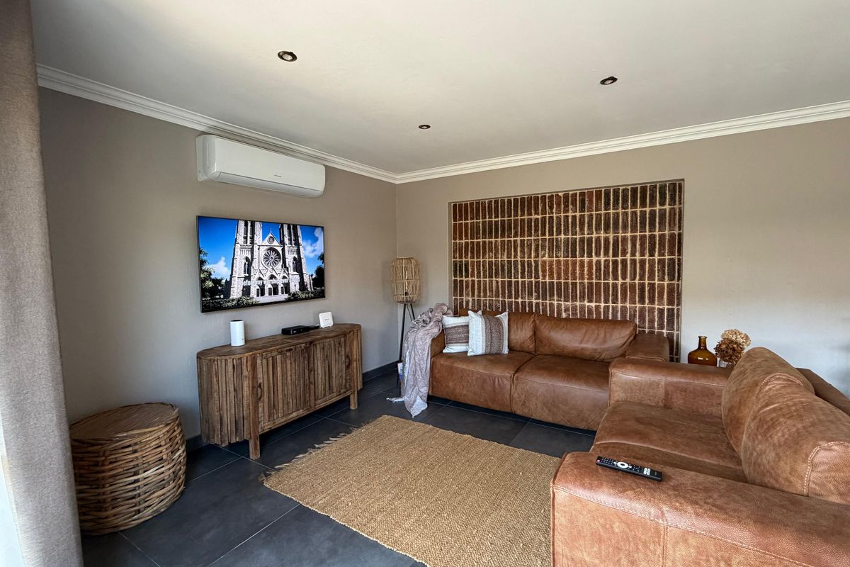 A well-maintained living room featuring a distinctive exposed brick accent wall, modern tiled flooring, recessed lighting, and a wall-mounted air conditioning unit.