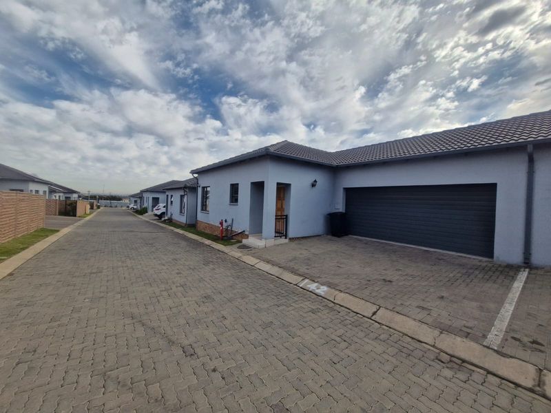 Front elevation of a modern townhouse featuring a paved driveway, automated garage door, and well-maintained plastered exterior walls within a residential complex.
