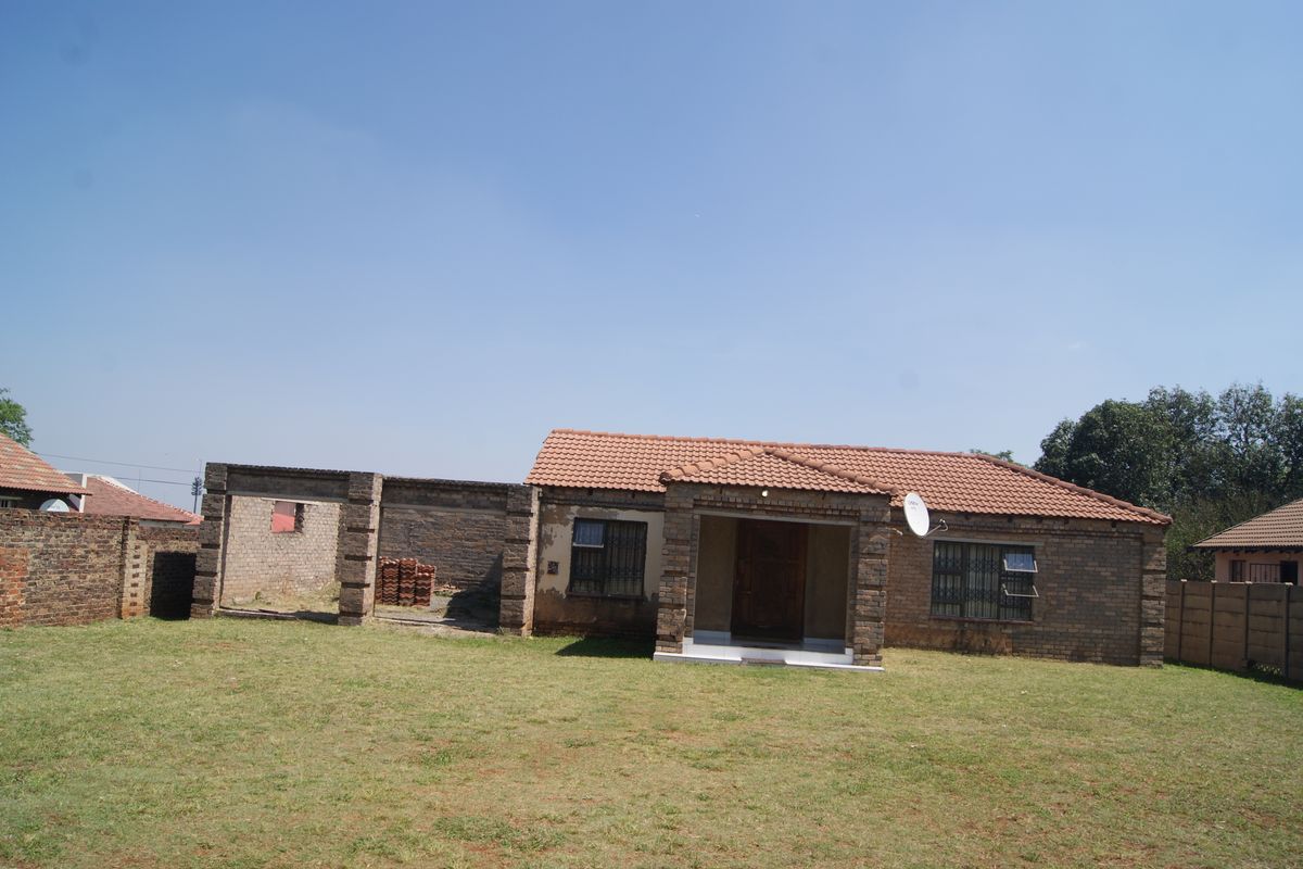 Rear view of a face brick house featuring a tiled roof, security bars, and an adjacent unfinished brick structure set within a large garden.