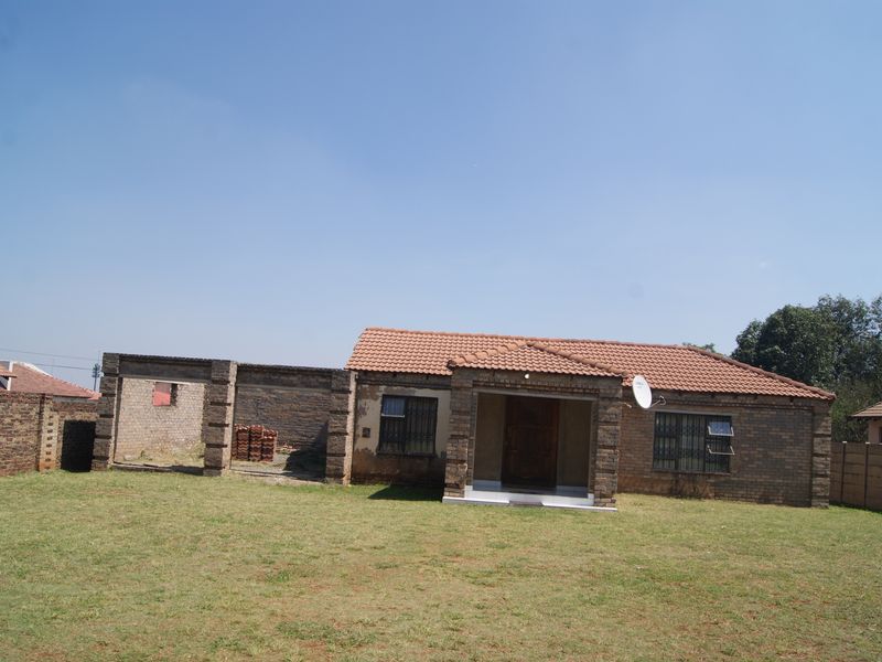 Rear view of a face brick house featuring a tiled roof, security bars, and an adjacent unfinished brick structure set within a large garden.