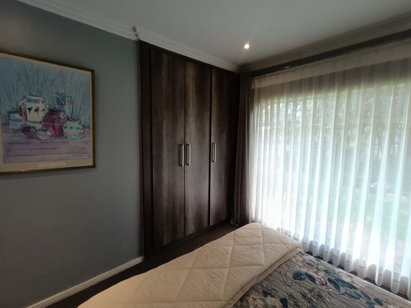 A well-maintained bedroom featuring dark wood built-in cupboards, recessed downlighting, and large windows with sheer curtains overlooking the garden.