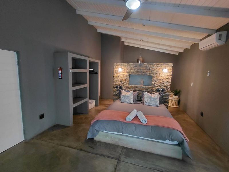 A spacious bedroom featuring a rustic stone accent wall, polished concrete floors, exposed timber ceiling beams, and integrated built-in storage shelving.