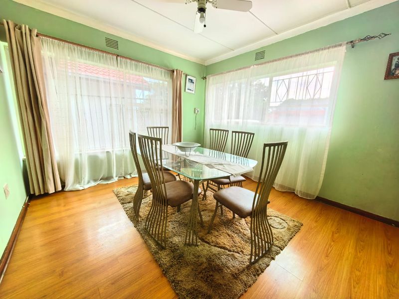A bright dining room featuring laminate flooring, a ceiling fan, and large windows with curtain rails, offering a functional space for family meals.