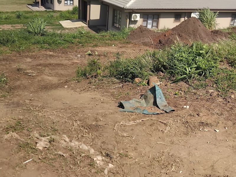 A view of a vacant residential plot featuring sloped, unimproved soil with adjacent houses visible in the background.