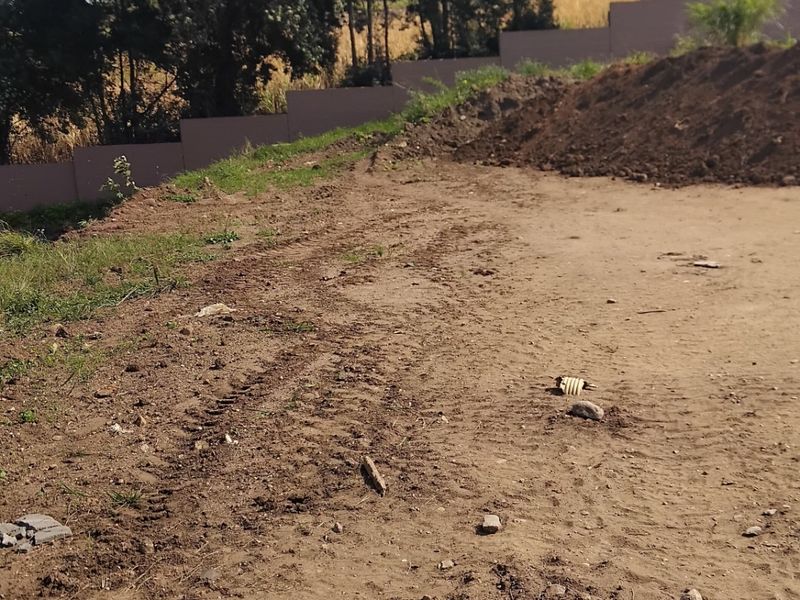 A vacant residential plot featuring cleared earth, tire tracks, and a perimeter boundary wall with mature trees in the background.