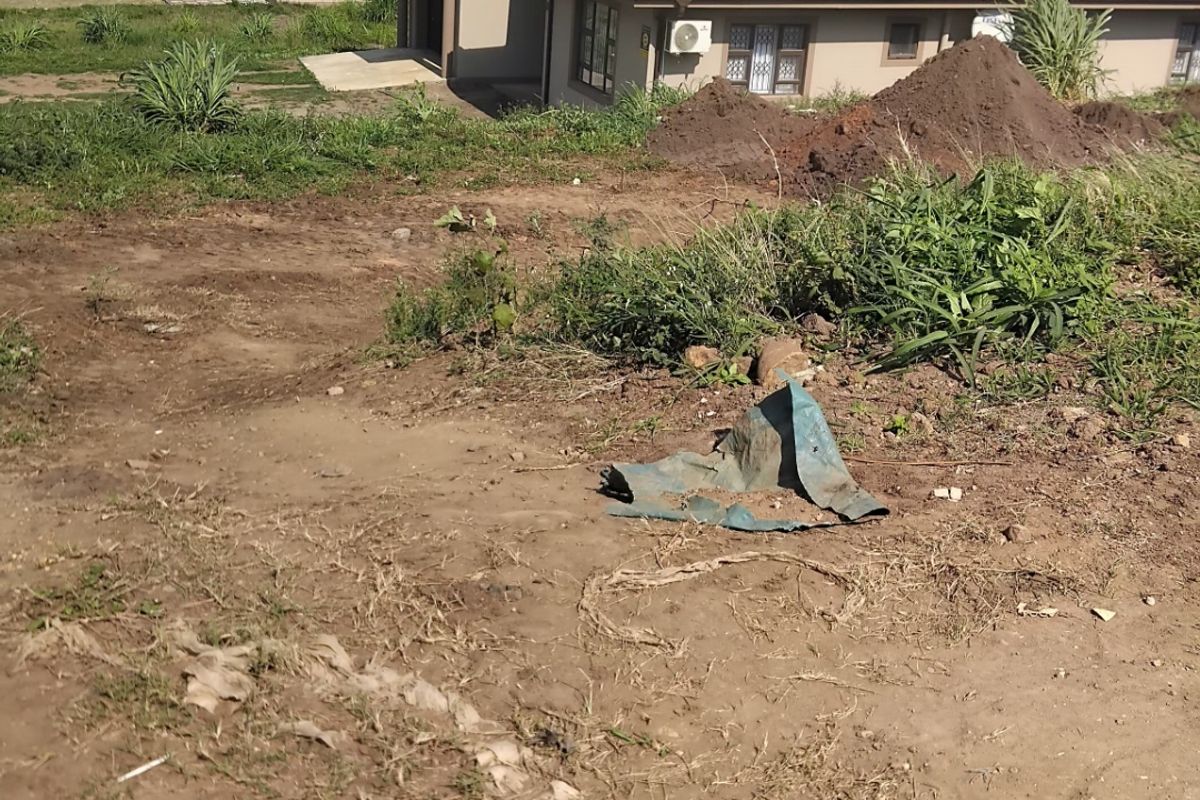 A view of a vacant residential plot featuring sloped, unimproved soil with adjacent houses visible in the background.