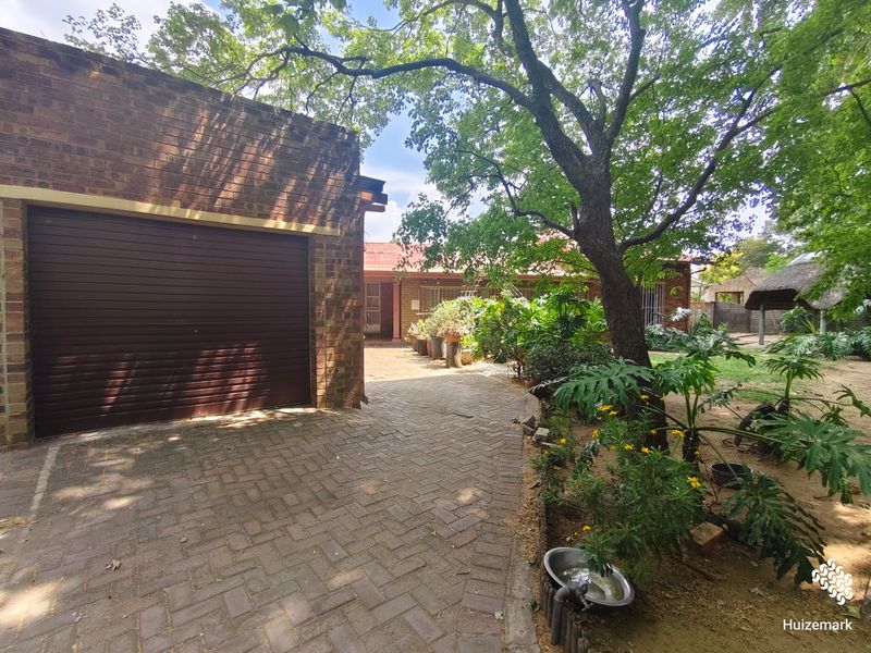 Front view of the property featuring a brick driveway, a single roller garage door, and established garden landscaping with mature trees.