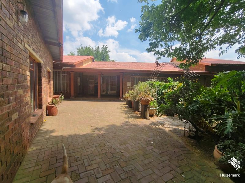 A spacious rear courtyard featuring brick paving, a covered patio area, and established garden beds against a classic brick house exterior.