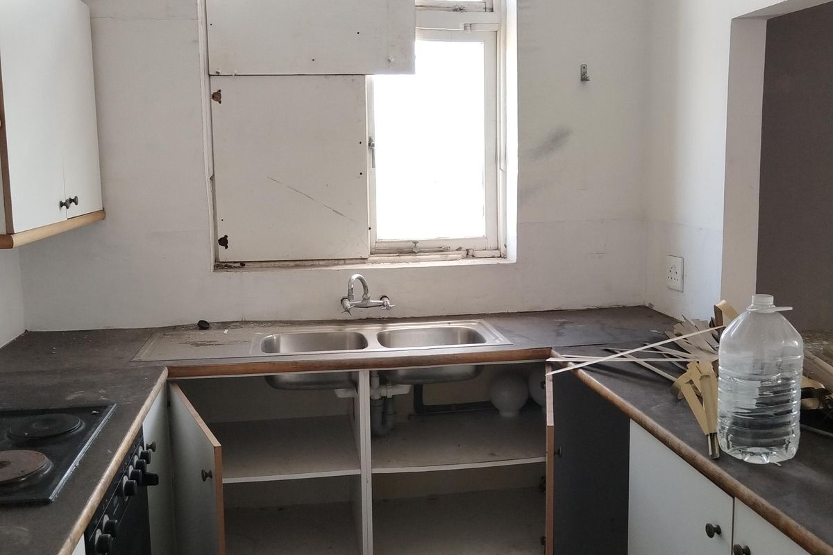 A dated kitchen featuring a double bowl sink, built-in oven and hob, and base cabinetry, requiring significant renovation and repair to the window and finishes.