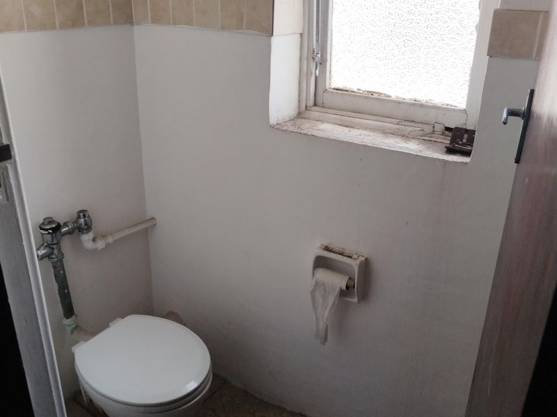 A small, dated guest toilet featuring tiled flooring, a wall-mounted flush valve toilet, and an obscured glass window requiring maintenance and cosmetic updates.